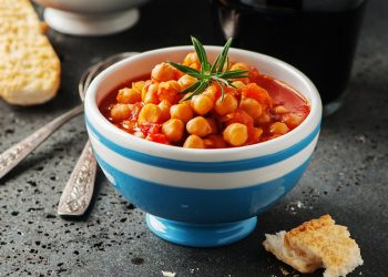 su tavolo nero un ciottola blu e bianca con una zuppa di ceci e rosmarino con accanto dei crostini e dietro 2 cucchiai
