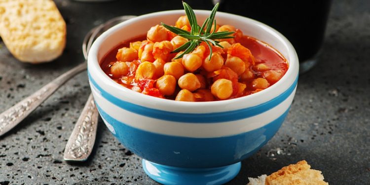 su tavolo nero un ciottola blu e bianca con una zuppa di ceci e rosmarino con accanto dei crostini e dietro 2 cucchiai