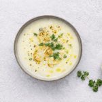 In primo piano su un tavolo chiaro c'è una zuppa di cavolfiore condita con olio EVO e del prezzemolo riccio.