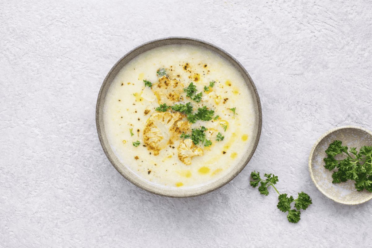 In primo piano su un tavolo chiaro c'è una zuppa di cavolfiore condita con olio EVO e del prezzemolo riccio.