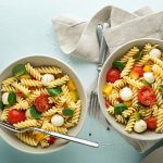 su sfondo verde chiaro 2 ciotole grandi con pasta pomodori e mozzarella e basilico