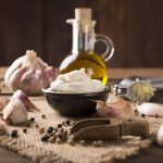 su tavolo di legno, sottopiatto in Juta con ciotola di terracotta con la salsa aioli e ingredienti come aglio, olio d'oliva e pepe