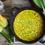 su base di legno una padella tonda con i manici e dentro una frittata di zucchine, accanto due zucchine col fiore