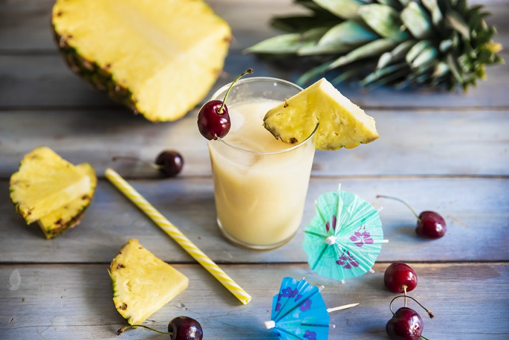 su base di legno un bicchiere con il cocktail Virgin Colada, spicchi di ananas e ciliegie