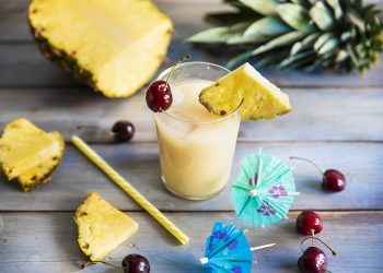 su base di legno un bicchiere con il cocktail Virgin Colada, spicchi di ananas e ciliegie