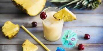 su base di legno un bicchiere con il cocktail Virgin Colada, spicchi di ananas e ciliegie