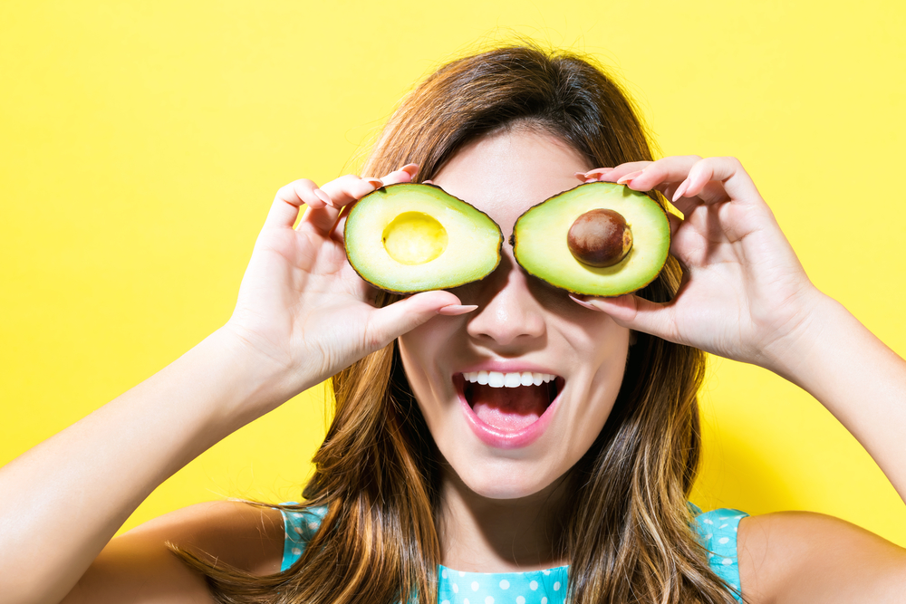 Su sfondo giallo, donna sorridente che ha messo sugli occhi un avocado per spiegare alimenti ricchi di potassio