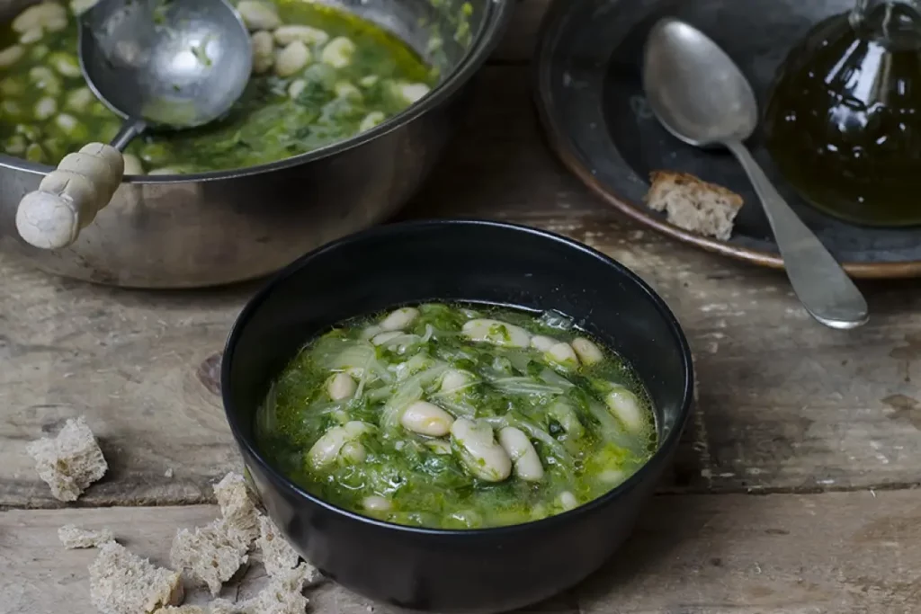 Scarola e fagioli: video e ricetta di una zuppa nutriente e sana 1 Su tavolo di legno, pentola con zuppa di scarola e fagioli con un mestolo. Accanto piatto di zuppa pronta con cucchiaio