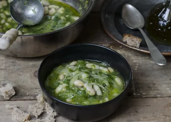 Su tavolo di legno, pentola con zuppa di scarola e fagioli con un mestolo. Accanto piatto di zuppa pronta con cucchiaio