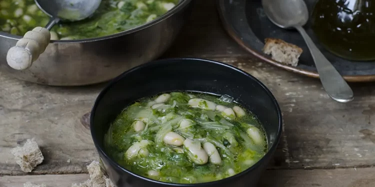 Su tavolo di legno, pentola con zuppa di scarola e fagioli con un mestolo. Accanto piatto di zuppa pronta con cucchiaio