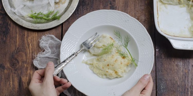 mani tengono una scodella bianca con finocchi gratinati, forchetta