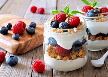 ricette con yogurt: due vasetti di vetro pieno di yogurt e frutti di bosco, un vassoio di legno con mirtilli e lamponi sparsi