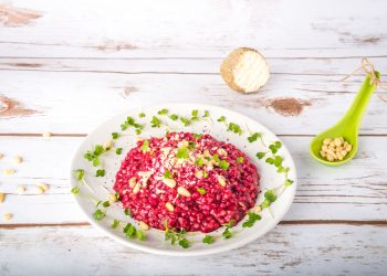 su tavolo di legno bianco un piatto bianco con un risotto alla barbabietola rossa decorato con semi e frutta secca. Accanto cucchiaio verde con pinoli