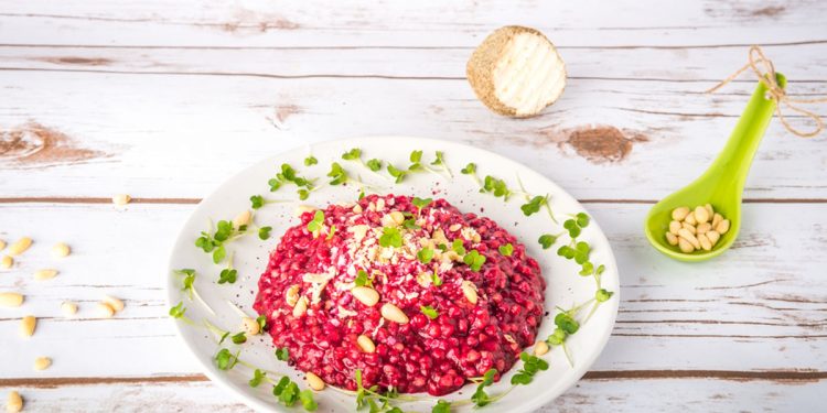 su tavolo di legno bianco un piatto bianco con un risotto alla barbabietola rossa decorato con semi e frutta secca. Accanto cucchiaio verde con pinoli