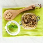 Pesto di melanzane cremoso e leggero 1 In primo piano su un canovaccio verde c'รจ un barattolo di vetro con del pesto di melanzane e una foglia di basilico sullo sfondo un mestolo di legno con dei pinoli.