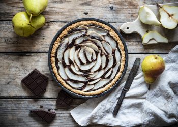 teglia con crostata pere e cioccolato, accanto a pere intere, pere tagliate, pezzi cioccolato, un coltello e un canovaccio