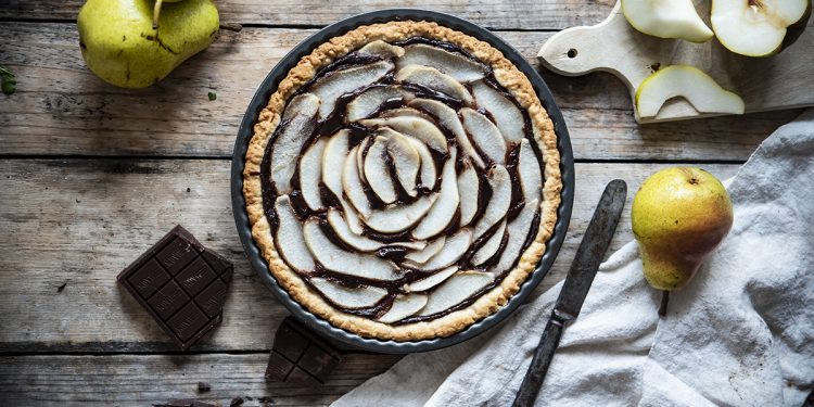 teglia con crostata pere e cioccolato, accanto a pere intere, pere tagliate, pezzi cioccolato, un coltello e un canovaccio