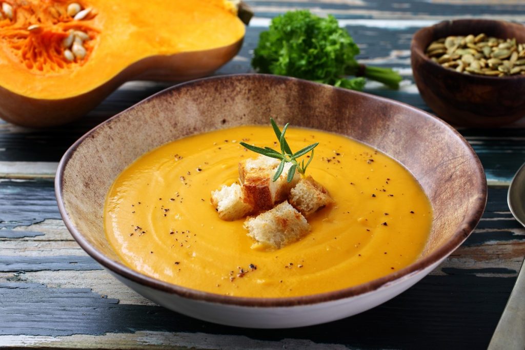 Vellutata di zucca, la ricetta gustosa e leggera 2 zuppiera rotonda con vellutata di zucca e alcuni crostini al centro, mezza zucca sullo sfondo
