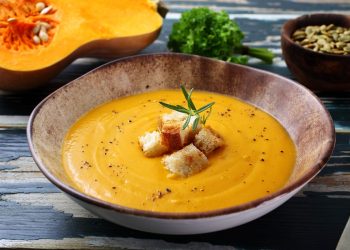 zuppiera rotonda con vellutata di zucca e alcuni crostini al centro, mezza zucca sullo sfondo
