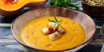 zuppiera rotonda con vellutata di zucca e alcuni crostini al centro, mezza zucca sullo sfondo