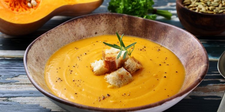 zuppiera rotonda con vellutata di zucca e alcuni crostini al centro, mezza zucca sullo sfondo