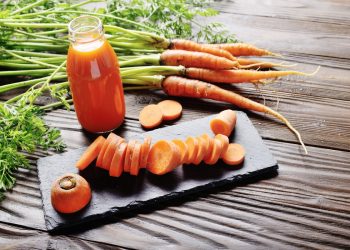 su base di legno, tagliere rettangolare con una carota cruda affettata a rondelle. Una bottiglia con del succo di carota e carote selvatiche intorno