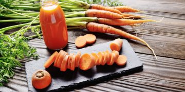 su base di legno, tagliere rettangolare con una carota cruda affettata a rondelle. Una bottiglia con del succo di carota e carote selvatiche intorno