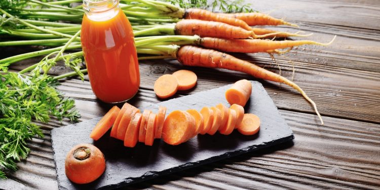 su base di legno, tagliere rettangolare con una carota cruda affettata a rondelle. Una bottiglia con del succo di carota e carote selvatiche intorno