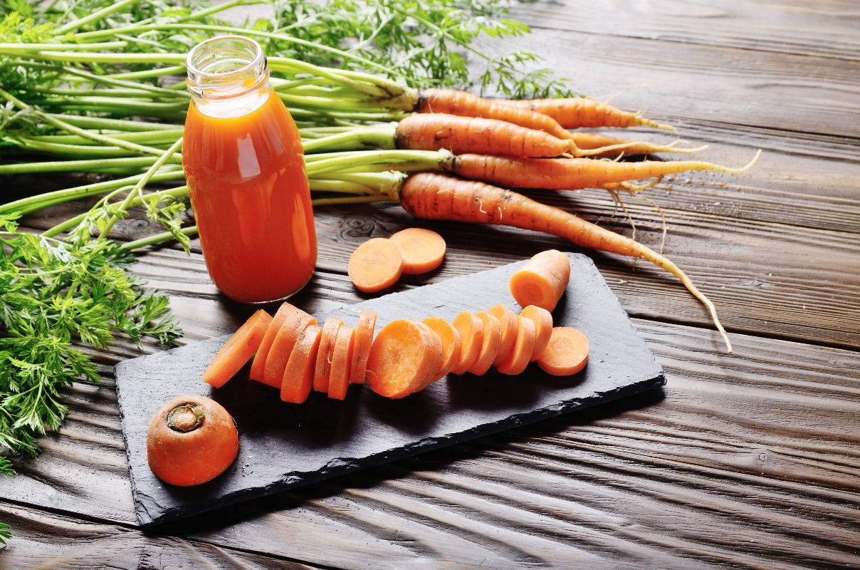 su base di legno, tagliere rettangolare con una carota cruda affettata a rondelle. Una bottiglia con del succo di carota e carote selvatiche intorno