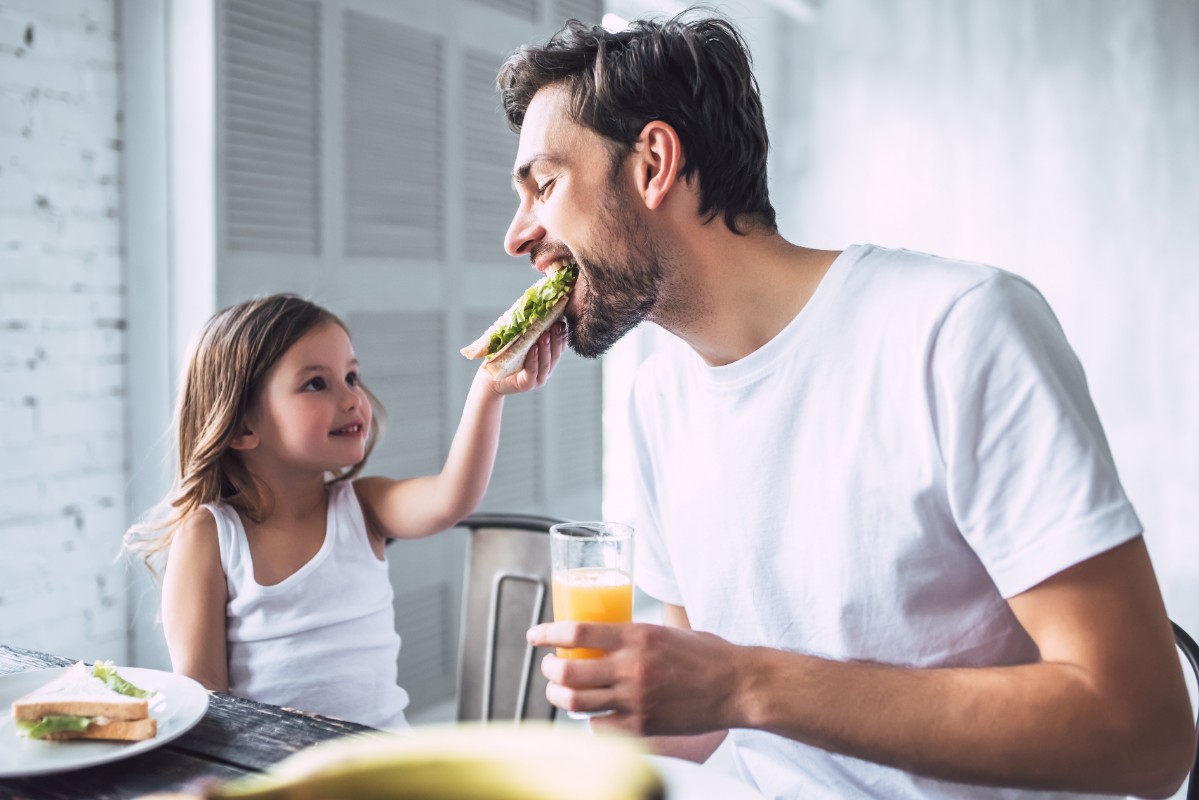 in una cucina, una bambina imbocca suo padre perché segue una dieta del panino. L'uomo tiene in mano una spremuta d'arancia. La bambina guarda il padre sorridendo e con tanto amore