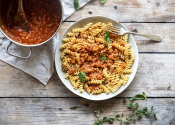 su base di legno un piatto chiaro pieno di fusilli con ragù di tempeh, accanto un pentolino con salsa di ragù