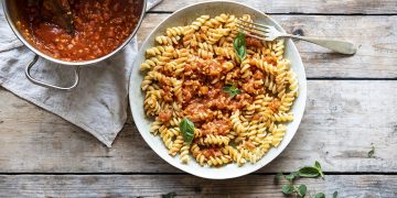 su base di legno un piatto chiaro pieno di fusilli con ragù di tempeh, accanto un pentolino con salsa di ragù
