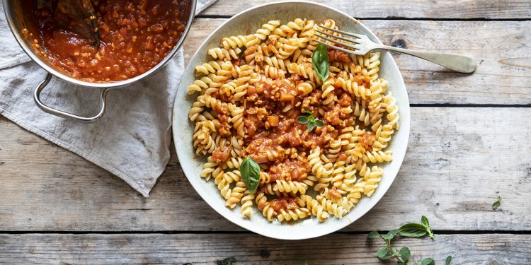 su base di legno un piatto chiaro pieno di fusilli con ragù di tempeh, accanto un pentolino con salsa di ragù