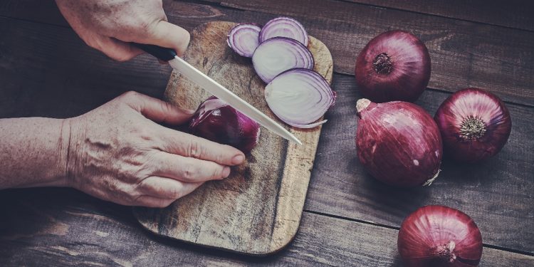 Due mani in primo piano che affettano una cipolla rossa di tropea su un tagliere