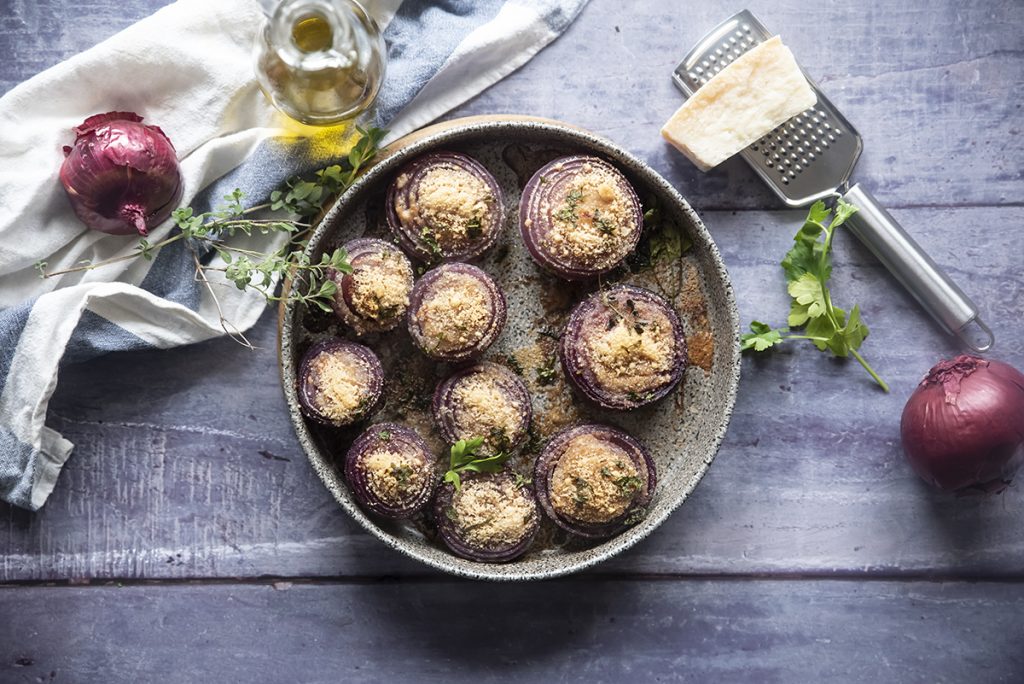 cipolle gratinate: su base di legno teglia tonda con cipolle gratinate, intorno: cipolle rosse, olio e parmigiano con grattugia
