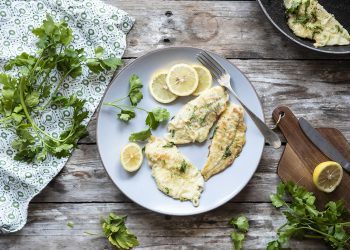 su tavolo di legno un piatto bianco con platessa alla mugnaia, erbe aromatiche attorno e una padella con un altra fetta di platessa
