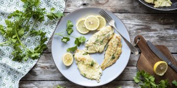 su tavolo di legno un piatto bianco con platessa alla mugnaia, erbe aromatiche attorno e una padella con un altra fetta di platessa