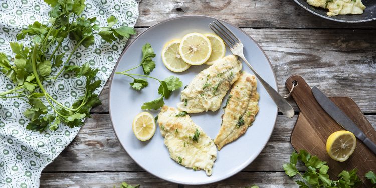 su tavolo di legno un piatto bianco con platessa alla mugnaia, erbe aromatiche attorno e una padella con un altra fetta di platessa