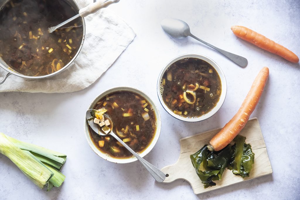 Zuppa di miso, la ricetta passo passo per farla in casa 4 su tavolo di marmo 2 contenitori bianchi con la zuppa di miso e attorno gli ingredienti: carota ,1 porro 2 cucchiai, e dietro la zuppiera piena di zuppa