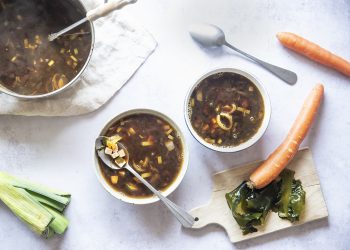 su tavolo di marmo 2 contenitori bianchi con la zuppa di miso e attorno gli ingredienti: carota ,1 porro 2 cucchiai, e dietro la zuppiera piena di zuppa