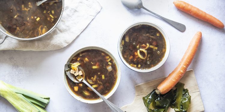 su tavolo di marmo 2 contenitori bianchi con la zuppa di miso e attorno gli ingredienti: carota ,1 porro 2 cucchiai, e dietro la zuppiera piena di zuppa
