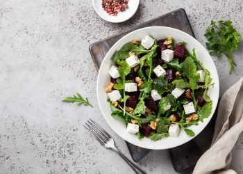 su piano di marmo con sotto tagliere nero piatto bianco con rucola, fetta e frutta secca. Accanto peperoncino e forchetta vicino al piatto
