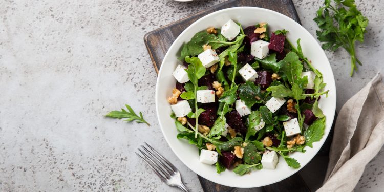 su piano di marmo con sotto tagliere nero piatto bianco con rucola, fetta e frutta secca. Accanto peperoncino e forchetta vicino al piatto