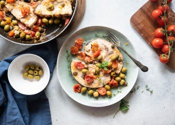 su tavolo bianco di marmo piatto verde con pesce alla siciliana e ciotola con olive verdi e pomodorini su tagliere di legno
