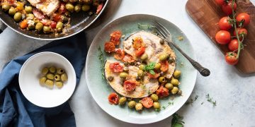 su tavolo bianco di marmo piatto verde con pesce alla siciliana e ciotola con olive verdi e pomodorini su tagliere di legno