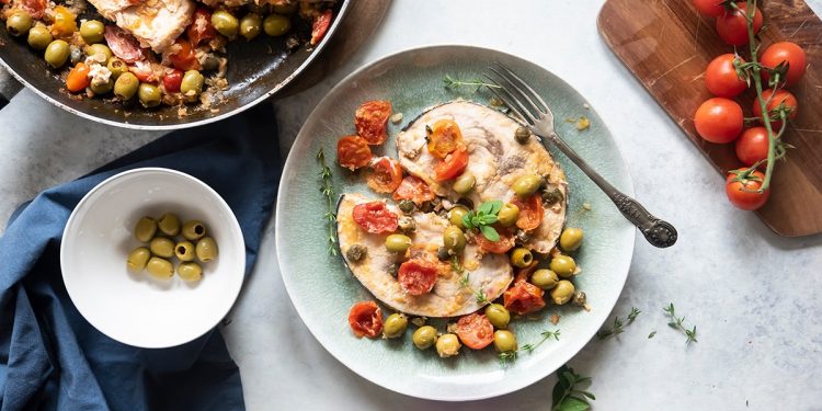 su tavolo bianco di marmo piatto verde con pesce alla siciliana e ciottola con olive verdi e pomodorini su tagliere di legno