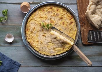 Su un tavolo di legno, un gateau di patate appena uscito dal forno con coltello e un uovo rotto