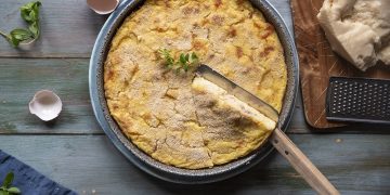 Su un tavolo di legno, un gateau di patate appena uscito dal forno con coltello e un uovo rotto