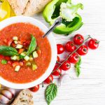 su tavolo di legno bianco, una scodella con dentro il gazpacho, attorno pomodorini, peperone verde limone e una bottiglia di olio