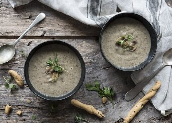 due ciotole scure con la vellutata di funghi, cucchiai e grissini sparsi su tavolo di legno